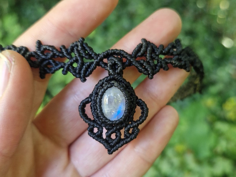 May include: Black macrame choker necklace featuring a central pendant with a pear-shaped, iridescent moonstone. The intricate knotwork forms a decorative pattern around the stone and along the choker's band. A handmade jewelry item.