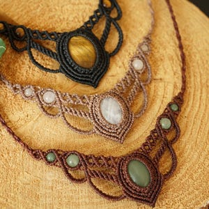 May include: Three handmade macrame necklaces with gemstone pendants. The top necklace is black with a yellow-brown stone, the middle is tan with a white stone, and the bottom is brown with a green stone. The necklaces are displayed on a wooden surface.