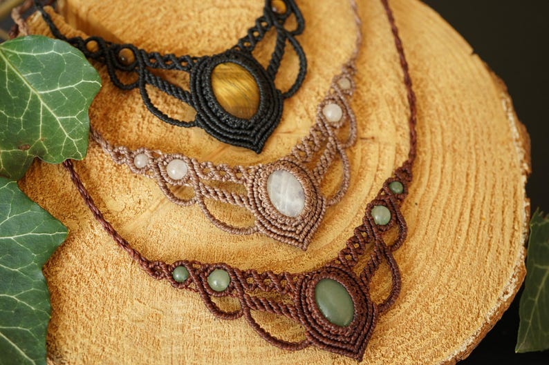 May include: Three handmade macrame necklaces with gemstone pendants. The top necklace is black with a yellow-brown stone, the middle is tan with a white stone, and the bottom is brown with a green stone. The necklaces are displayed on a wooden surface.
