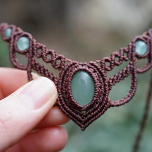 May include: A handmade macrame necklace featuring a brown cord design with a central oval-shaped green stone and smaller green stones. The intricate woven pattern creates a unique, bohemian style.