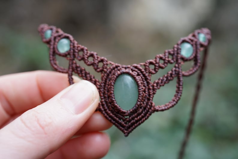 May include: A handmade macrame necklace featuring a brown cord design with a central oval-shaped green stone and smaller green stones. The intricate woven pattern creates a unique, bohemian style.