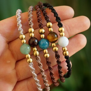 May include: Five handmade macrame bracelets with gemstone beads in various colors, including green, brown, blue, and white. Each bracelet features gold-colored beads and a knotted cord. The bracelets are displayed against a blurred green background.