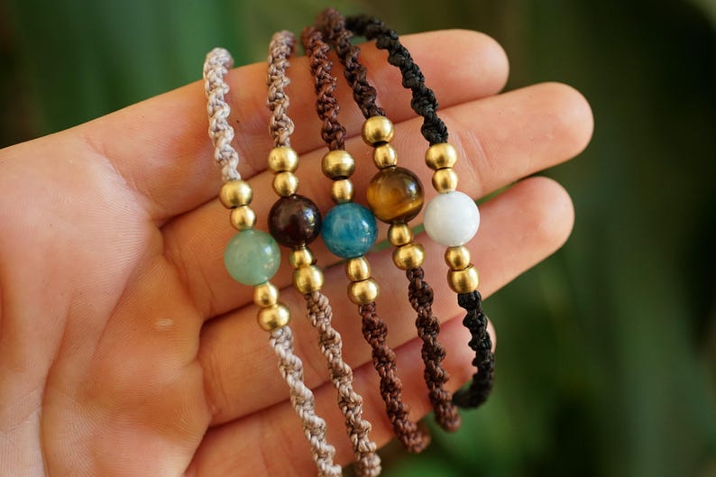 May include: Five handmade macrame bracelets with gemstone beads in various colors, including green, brown, blue, and white. Each bracelet features gold-colored beads and a knotted cord. The bracelets are displayed against a blurred green background.