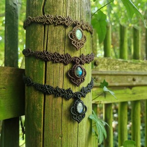 May include: Three macrame chokers with gemstone pendants are displayed on a weathered wooden post. The chokers are in shades of brown and black, each featuring a central pendant with a blue-green stone. The background is a blurred view of greenery.