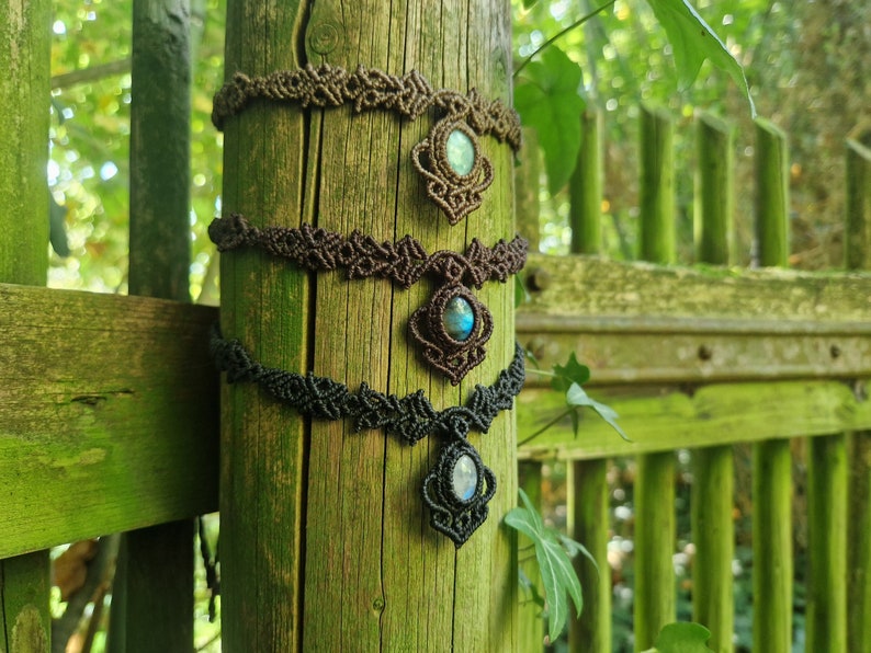 May include: Three macrame chokers with gemstone pendants are displayed on a weathered wooden post. The chokers are in shades of brown and black, each featuring a central pendant with a blue-green stone. The background is a blurred view of greenery.