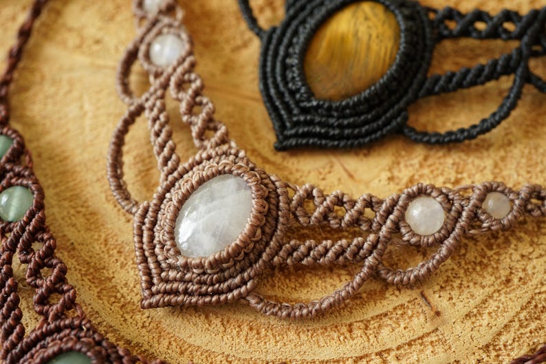 May include: Close-up of three macrame necklaces. The necklaces are brown and black, with oval and round gemstones. The brown necklace has a large oval gemstone in the center and smaller round gemstones. The black necklace has a large oval gemstone.