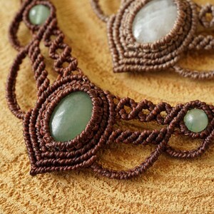 May include: Close-up of two macrame necklaces. The necklaces are made of brown cord and feature oval-shaped green gemstones. The macrame work is intricate, with detailed knots and patterns. The necklaces are displayed on a light brown wooden surface.