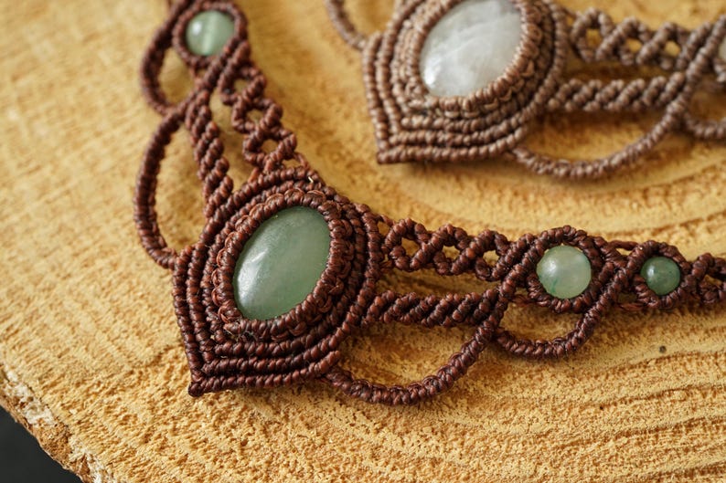 May include: Close-up of two macrame necklaces. The necklaces are made of brown cord and feature oval-shaped green gemstones. The macrame work is intricate, with detailed knots and patterns. The necklaces are displayed on a light brown wooden surface.