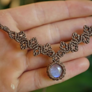 Puede incluir: Un collar de macram&eacute; marr&oacute;n con una piedra cabuj&oacute;n circular central que presenta un brillo iridiscente azul p&aacute;lido. La banda del collar est&aacute; elaborada con cord&oacute;n anudado intrincado, formando un patr&oacute;n geom&eacute;trico repetitivo desde la piedra.