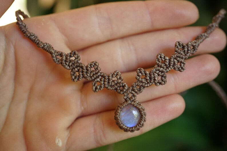 Puede incluir: Un collar de macram&eacute; marr&oacute;n con una piedra cabuj&oacute;n circular central que presenta un brillo iridiscente azul p&aacute;lido. La banda del collar est&aacute; elaborada con cord&oacute;n anudado intrincado, formando un patr&oacute;n geom&eacute;trico repetitivo desde la piedra.