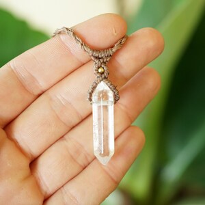 May include: A clear, hexagonal, double-terminated quartz crystal pendant suspended from a brown macrame cord. The crystal is secured by intricate brown fiber knotting and features a small, round, metallic gold-colored bead accent at the top.