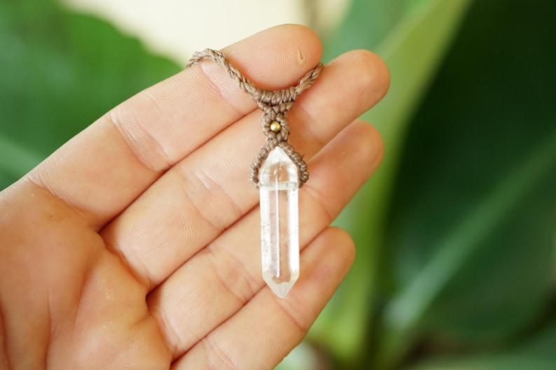 May include: A clear, hexagonal, double-terminated quartz crystal pendant suspended from a brown macrame cord. The crystal is secured by intricate brown fiber knotting and features a small, round, metallic gold-colored bead accent at the top.