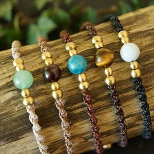 May include: Five handmade bracelets with gemstone beads and gold-colored accents. The bracelets feature a variety of colors, including green, brown, blue, and white, and are displayed on a piece of wood.