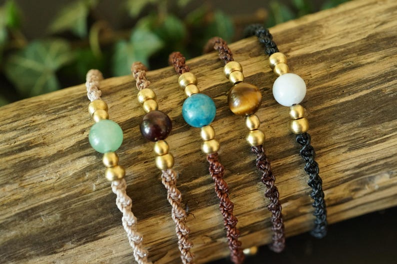 May include: Five handmade bracelets with gemstone beads and gold-colored accents. The bracelets feature a variety of colors, including green, brown, blue, and white, and are displayed on a piece of wood.