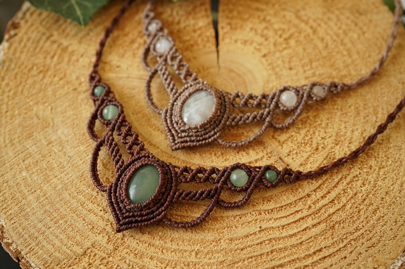 May include: Two handmade macrame necklaces with gemstone accents. One necklace features a large oval green stone and smaller green beads, while the other has a large oval white stone and smaller white beads. Both are brown.