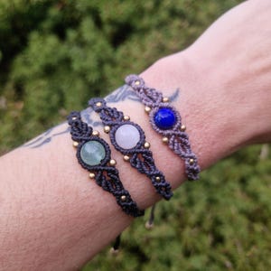 May include: Three macrame bracelets with gemstone accents. One bracelet features a green stone, another a white stone, and the third a blue stone. Each bracelet is approximately 0.75 inches wide and has gold-colored beads.