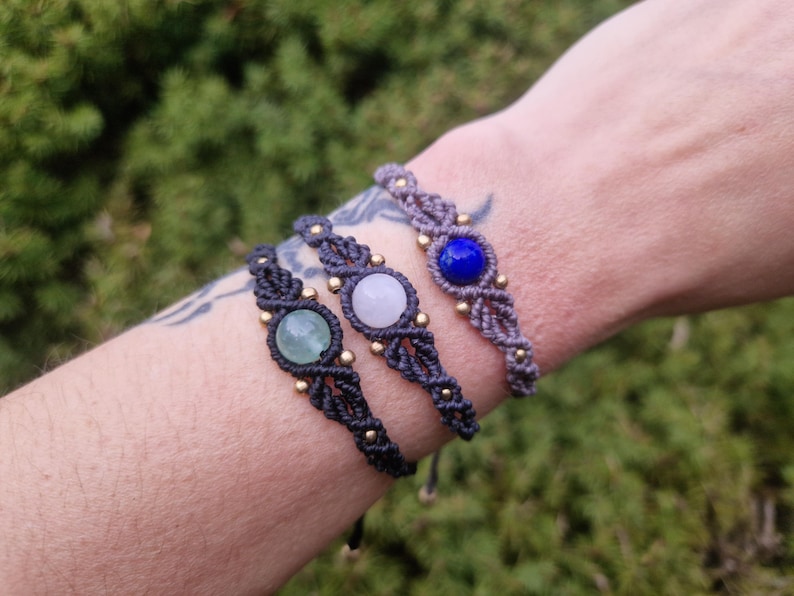 May include: Three macrame bracelets with gemstone accents. One bracelet features a green stone, another a white stone, and the third a blue stone. Each bracelet is approximately 0.75 inches wide and has gold-colored beads.