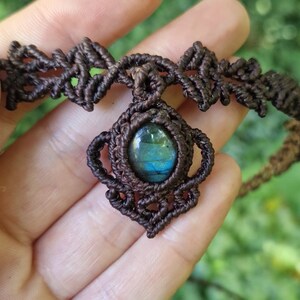 May include: A handcrafted macrame necklace featuring a central oval-shaped gemstone with blue and green hues. The dark brown macrame cord forms an intricate design around the stone and the necklace's band. The necklace is held in a hand against a blurred green background.
