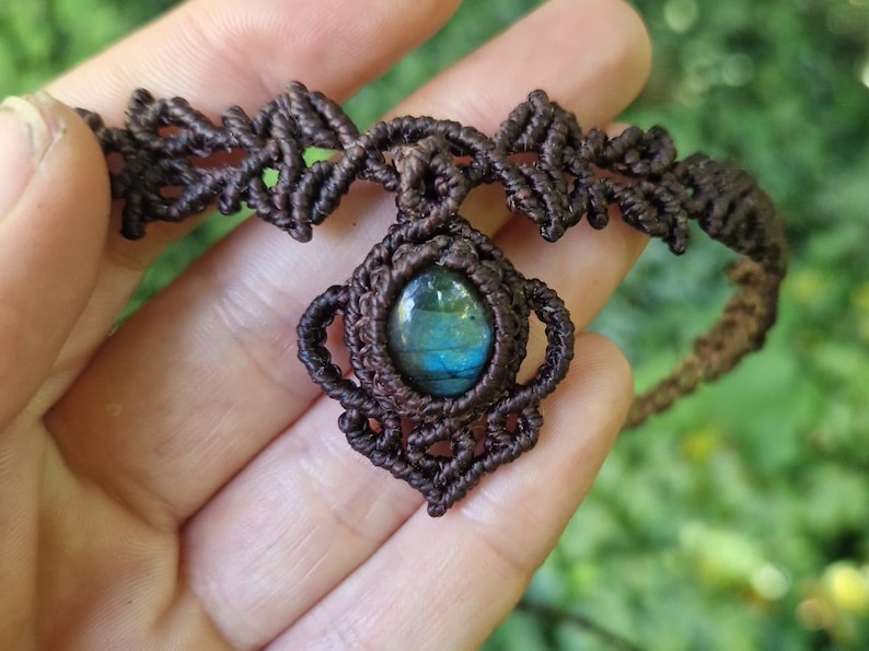 May include: A handcrafted macrame necklace featuring a central oval-shaped gemstone with blue and green hues. The dark brown macrame cord forms an intricate design around the stone and the necklace's band. The necklace is held in a hand against a blurred green background.