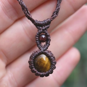 May include: A handcrafted macrame necklace featuring a tiger's eye gemstone pendant. The pendant is a warm, golden-brown color with dark brown accents, set in a woven, dark brown macrame design. A smaller, round, dark red bead is also incorporated.