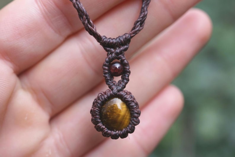 May include: A handcrafted macrame necklace featuring a tiger's eye gemstone pendant. The pendant is a warm, golden-brown color with dark brown accents, set in a woven, dark brown macrame design. A smaller, round, dark red bead is also incorporated.