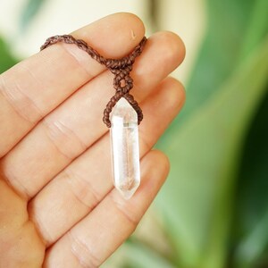 May include: A clear, double-terminated quartz crystal pendant suspended from a dark brown macrame cord. The crystal is faceted into a hexagonal point shape and is secured by a knotted brown cord design that wraps around the top of the stone.