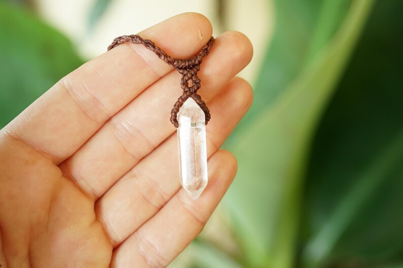 May include: A clear, double-terminated quartz crystal pendant suspended from a dark brown macrame cord. The crystal is faceted into a hexagonal point shape and is secured by a knotted brown cord design that wraps around the top of the stone.