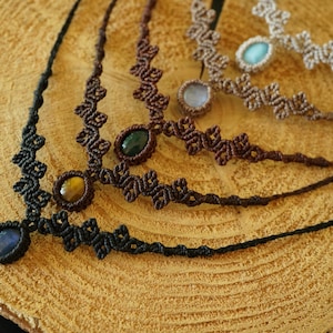 Puede incluir: Una colecci&oacute;n de cuatro collares de macram&eacute; con dise&ntilde;os de cord&oacute;n anudado en negro, marr&oacute;n oscuro y beige. Cada collar est&aacute; centrado con una piedra preciosa ovalada, incluyendo piedras azules, ojo de tigre, verde oscuro y azul claro.