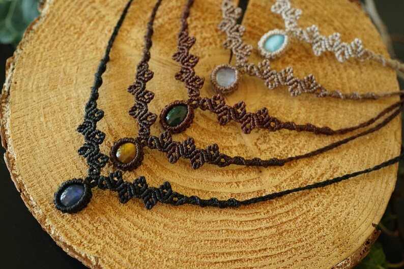 Puede incluir: Una colecci&oacute;n de cuatro collares de macram&eacute; con dise&ntilde;os de cord&oacute;n anudado en negro, marr&oacute;n oscuro y beige. Cada collar est&aacute; centrado con una piedra preciosa ovalada, incluyendo piedras azules, ojo de tigre, verde oscuro y azul claro.