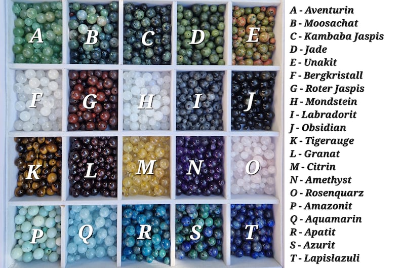 May include: A collection of gemstone beads in a white divided tray. The beads are in various colors, including green, brown, white, blue, and purple. Each section is labeled with a letter and a gemstone name, such as Aventurin, Jade, and Amethyst.