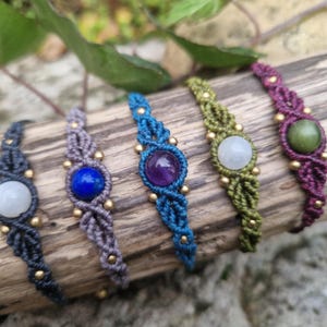 May include: A collection of macrame bracelets with various colored cords and gemstones. The bracelets are displayed on a piece of wood. Gemstones include green, white, blue, and purple. Gold-colored beads accent the macrame.