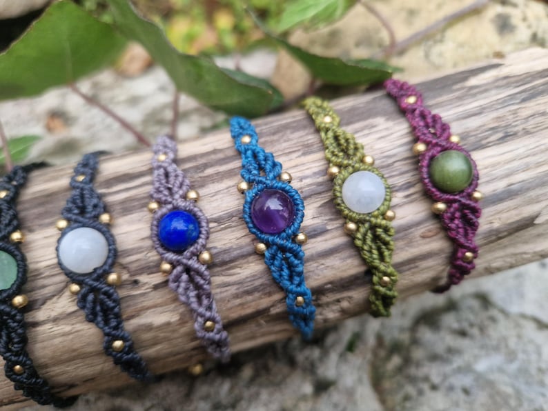 May include: A collection of macrame bracelets with various colored cords and gemstones. The bracelets are displayed on a piece of wood. Gemstones include green, white, blue, and purple. Gold-colored beads accent the macrame.
