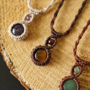 May include: Close-up of three handmade macrame necklaces with gemstone pendants. The necklaces feature brown, beige, and black cords, with round gemstone pendants in shades of purple, yellow, and green. The necklaces are displayed on a wooden surface.