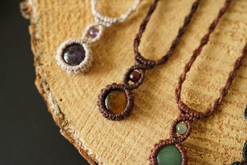 May include: Close-up of three handmade macrame necklaces with gemstone pendants. The necklaces feature brown, beige, and black cords, with round gemstone pendants in shades of purple, yellow, and green. The necklaces are displayed on a wooden surface.