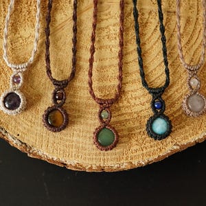 May include: A collection of handmade macrame necklaces with gemstone pendants. Each necklace features a different colored gemstone, including amethyst, tiger's eye, green aventurine, and rose quartz. The necklaces are displayed on a wooden surface.