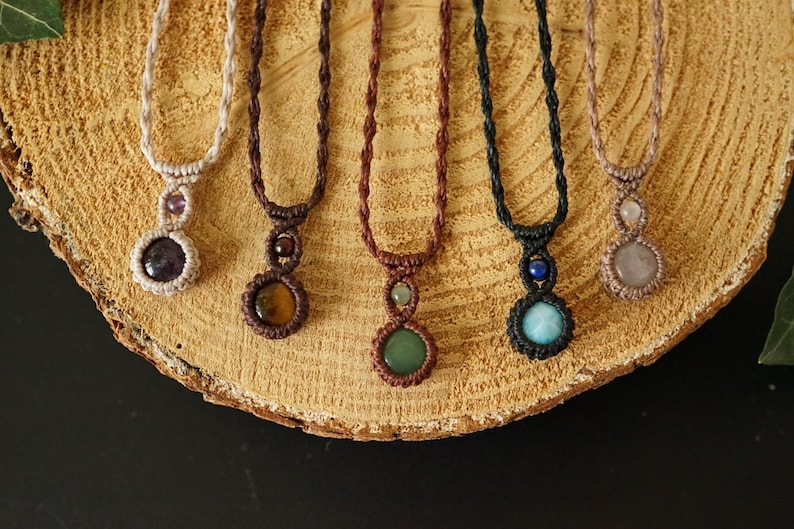 May include: A collection of handmade macrame necklaces with gemstone pendants. Each necklace features a different colored gemstone, including amethyst, tiger's eye, green aventurine, and rose quartz. The necklaces are displayed on a wooden surface.