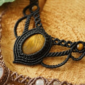 May include: Close-up of two macrame necklaces. The black necklace features a large oval tiger's eye stone and smaller beads. The second necklace is brown with white stones. Both necklaces are on a textured, light brown surface.