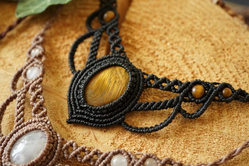 May include: Close-up of two macrame necklaces. The black necklace features a large oval tiger's eye stone and smaller beads. The second necklace is brown with white stones. Both necklaces are on a textured, light brown surface.