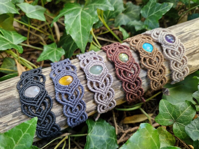 May include: A collection of six macrame bracelets with gemstone accents. The bracelets are in various colors, including black, gray, brown, and tan. Each bracelet features a different gemstone, such as quartz, tiger's eye, and turquoise.