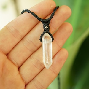 May include: A clear, pointed crystal pendant suspended from a black cord. The cord features intricate macrame knotting at the top of the crystal, accented by a small, round, metallic silver bead integrated into the woven black fiber design.