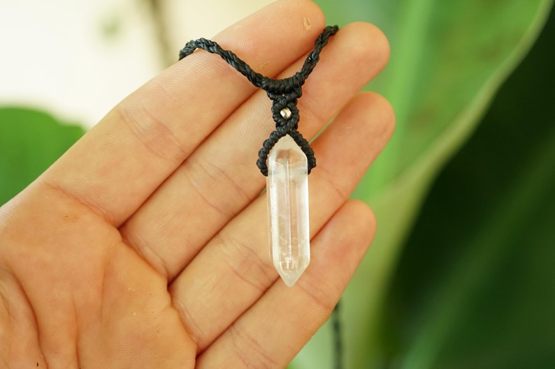 May include: A clear, pointed crystal pendant suspended from a black cord. The cord features intricate macrame knotting at the top of the crystal, accented by a small, round, metallic silver bead integrated into the woven black fiber design.