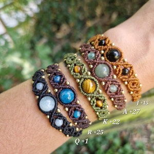 May include: A collection of macrame bracelets in various colors, including black, brown, and olive green. Each bracelet features different gemstones and gold-colored beads. The bracelets are displayed on a wrist.