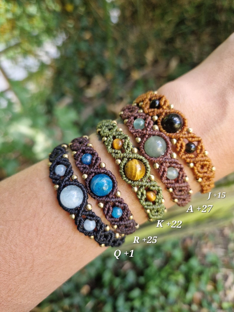 May include: A collection of macrame bracelets in various colors, including black, brown, and olive green. Each bracelet features different gemstones and gold-colored beads. The bracelets are displayed on a wrist.