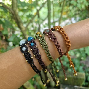 May include: A collection of macrame bracelets in various colors, including black, brown, green, and tan. Each bracelet features gemstone beads and gold-colored accents. The bracelets are displayed on a person's arm against a blurred green background.