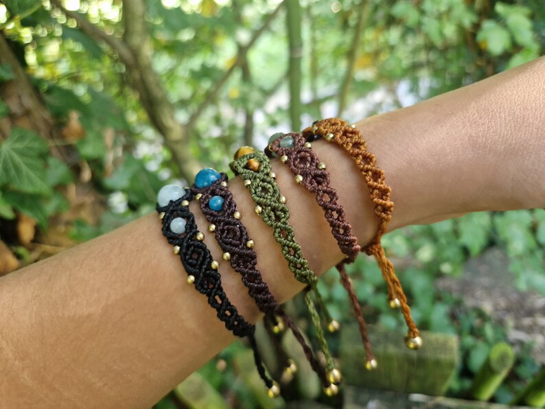 May include: A collection of macrame bracelets in various colors, including black, brown, green, and tan. Each bracelet features gemstone beads and gold-colored accents. The bracelets are displayed on a person's arm against a blurred green background.