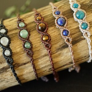 May include: Five handmade macrame bracelets with gemstone beads. The bracelets feature a variety of colors, including white, green, brown, blue, and turquoise. Each bracelet is displayed on a piece of driftwood.