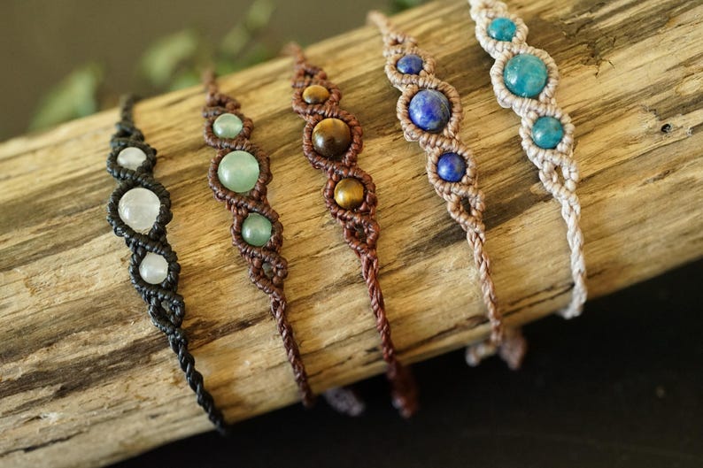 May include: Five handmade macrame bracelets with gemstone beads. The bracelets feature a variety of colors, including white, green, brown, blue, and turquoise. Each bracelet is displayed on a piece of driftwood.