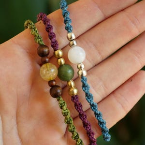 May include: Close-up of three handmade beaded bracelets. Each bracelet features a different color of knotted cord: green, purple, and blue. The bracelets have wooden, gold, and gemstone beads.