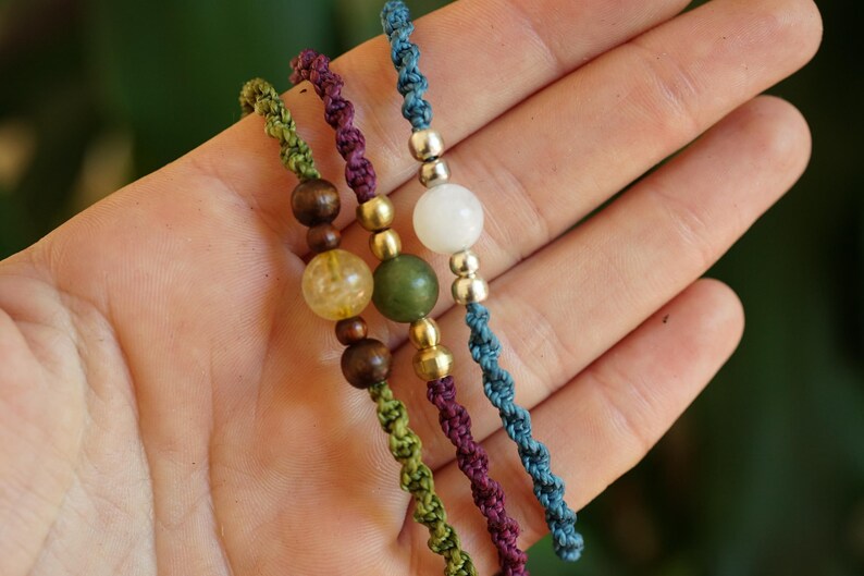 May include: Close-up of three handmade beaded bracelets. Each bracelet features a different color of knotted cord: green, purple, and blue. The bracelets have wooden, gold, and gemstone beads.