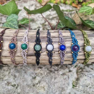 May include: A collection of eight macrame bracelets, each featuring a different gemstone. The bracelets are displayed on a piece of wood, showcasing a variety of colors including brown, blue, green, and purple. The gemstones are round and set in the center of each bracelet.